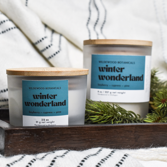 Two winter wonderland soy candles by Wildewood Botanicals on a wooden surface with a white and black striped fabric background.