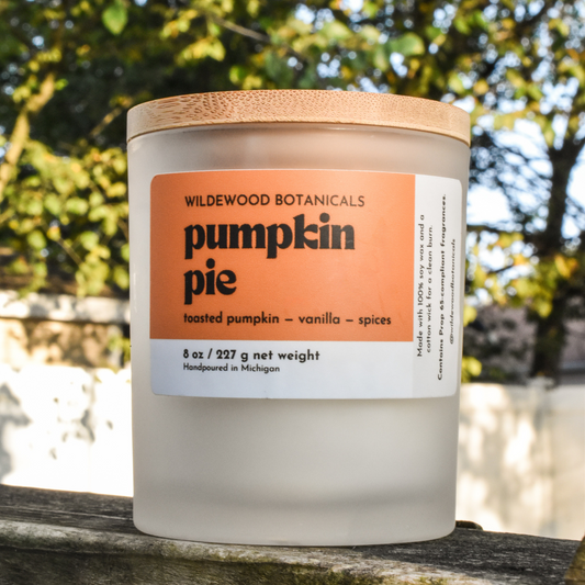 Candle jar labeled 'Pumpkin Pie' by Wildewood Botanicals on a wooden surface with a blurred natural background.