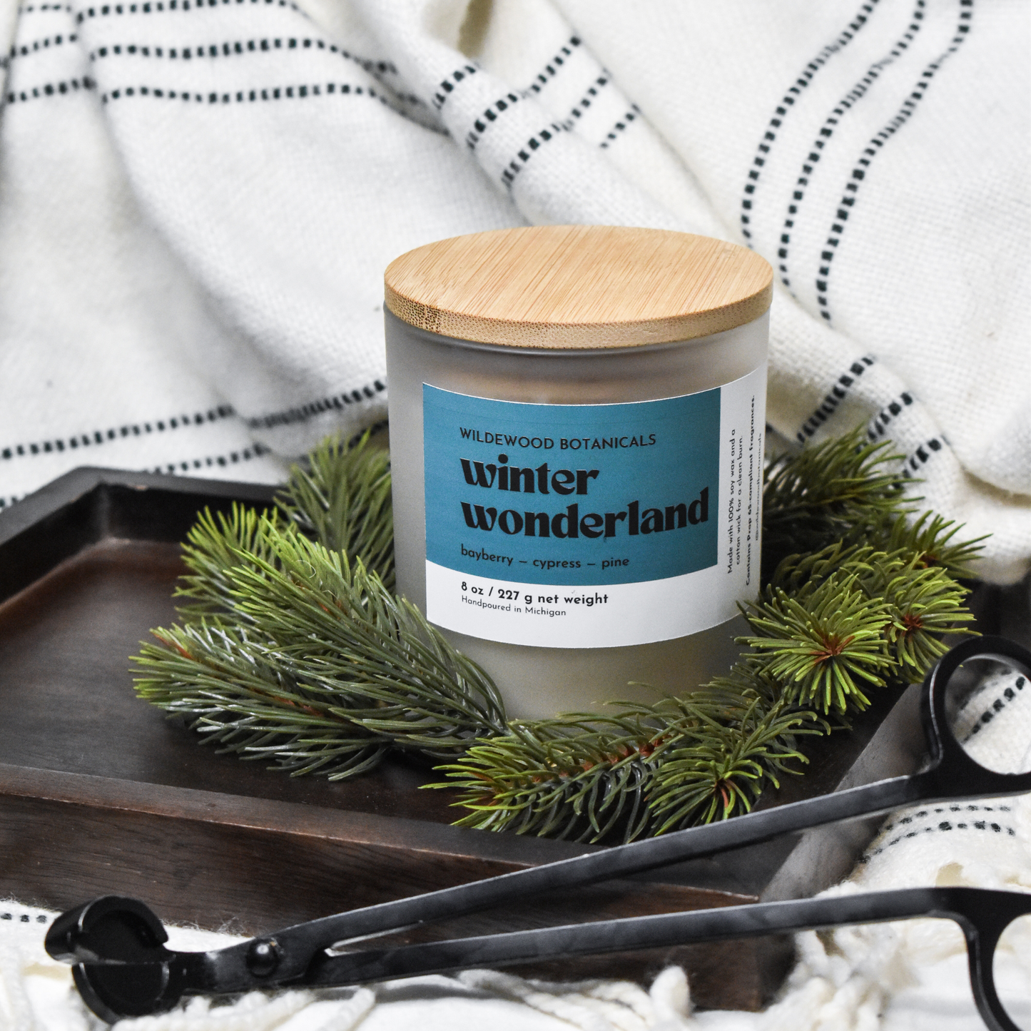 Soy candle labeled 'Winter Wonderland' with a wooden lid on a tray with greenery and a black tool.